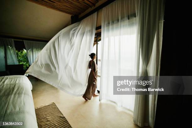 wide shot of woman standing in luxury hotel suite looking at view with curtains blowing in wind - raffiné photos et images de collection