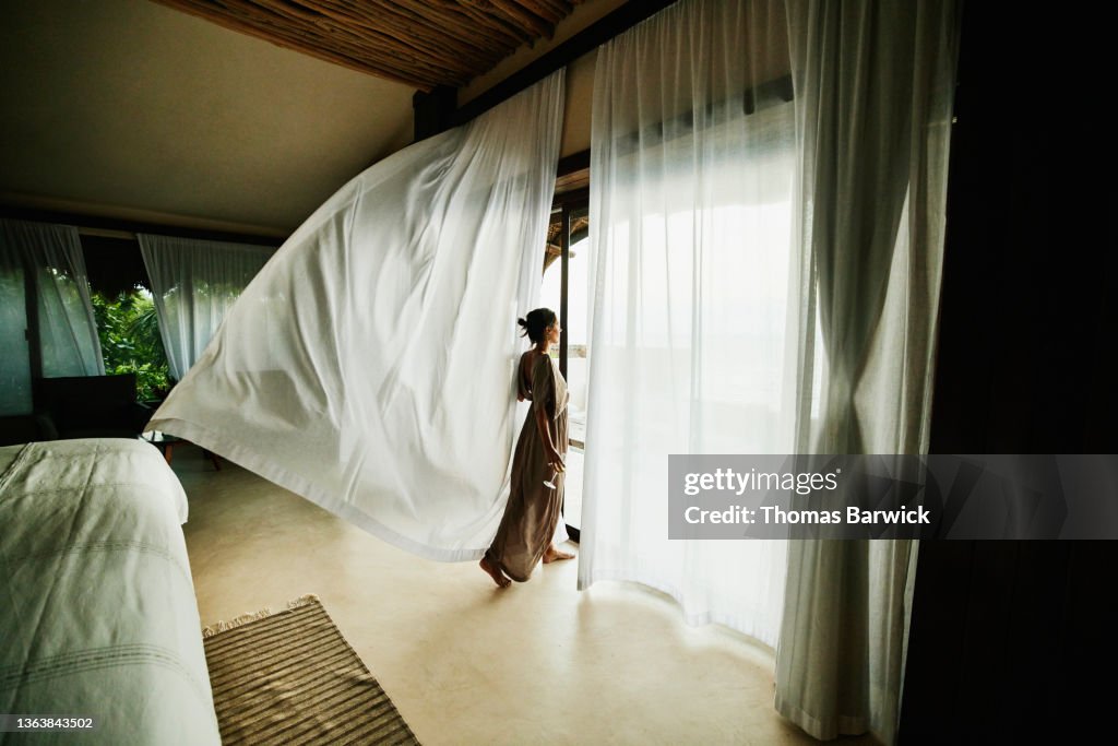 Wide shot of woman standing in luxury hotel suite looking at view with curtains blowing in wind