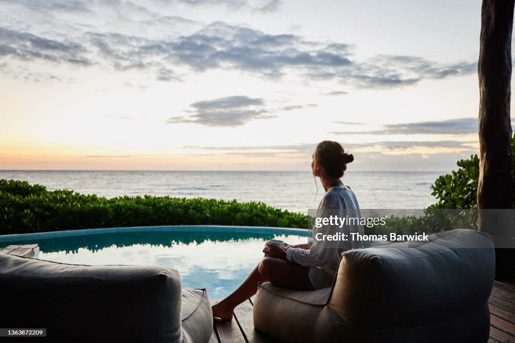 Wide shot of woman watching sunrise while sitting poolside at luxury suite at tropical resort
