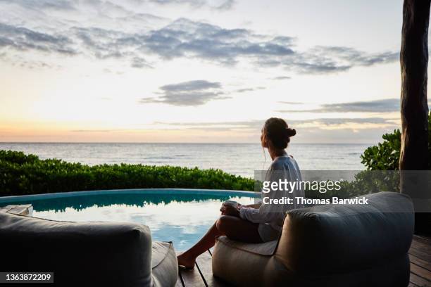 wide shot of woman watching sunrise while sitting poolside at luxury suite at tropical resort - frau anfang 30 stock-fotos und bilder