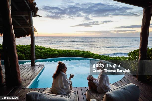 wide shot of female friends in discussion while sitting poolside at luxury suite of tropical resort at sunrise - luxury pool stock pictures, royalty-free photos & images