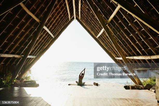 wide shot of woman warming up before practicing yoga in ocean front pavilion at tropical resort - inviting gesture stock pictures, royalty-free photos & images