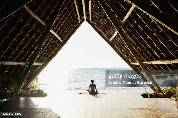 wide shot of woman relaxing after practicing yoga in ocean front pavilion at tropical resort - bienestar fotografías e imágenes de stock