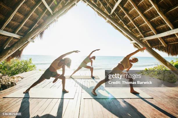 wide shot of women in extended side angle pose while practicing yoga during class in ocean front pavilion at tropical resort - hotel ecologico fotografías e imágenes de stock