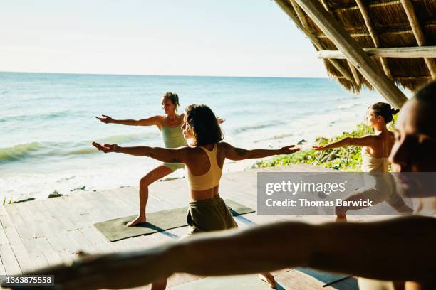 wide shot of women in warrior pose while practicing yoga during class in ocean front pavilion at tropical resort - group sea stock-fotos und bilder
