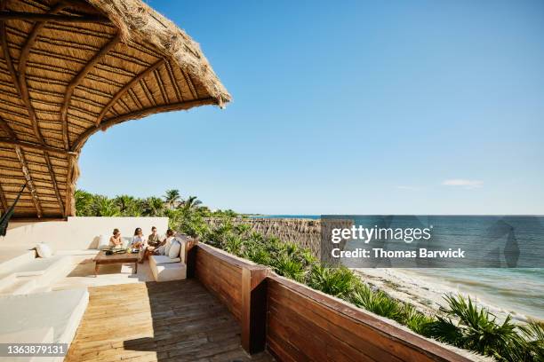 extreme wide shot of female friends sharing breakfast on deck of luxury suite overlooking ocean at tropical resort - dining overlooking water stock-fotos und bilder