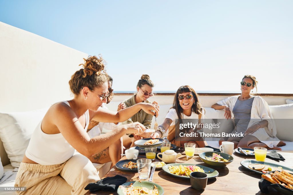 Medium wide shot of smiling female friends sharing breakfast on deck of luxury suite at tropical resort