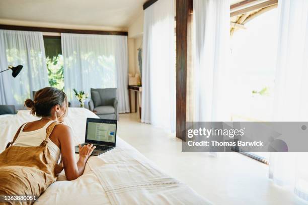 wide shot of woman working on laptop while relaxing on bed in luxury suite of tropical resort - hotel room work stock-fotos und bilder