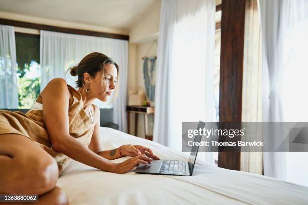 wide shot of woman working on laptop while relaxing on bed in luxury suite of tropical resort - cama lujo fotografías e imágenes de stock