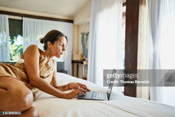 Wide shot of woman working on laptop while relaxing on bed in luxury suite of tropical resort