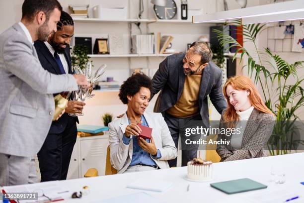 woman opening her gift box with colleagues standing by holding champagne glasses. - corporate gift policy stock pictures, royalty-free photos & images