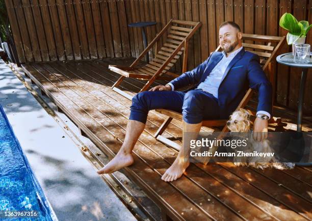 confident and successful businessman outdoors relaxing on a wooden folding chair next to the swimming pool and pampering his dog - well dressed dog stock pictures, royalty-free photos & images