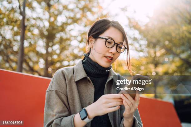 young asian woman using smartphone, sitting on the bench in urban park against sunlight and green plants. taking a break and enjoying the fresh air on beautiful autumn day - orange tree stock-fotos und bilder