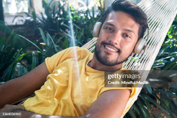 cheerful latin man looking at the camera while listening to music on headphones - young man listening music and relaxing in hammock stock pictures, royalty-free photos & images