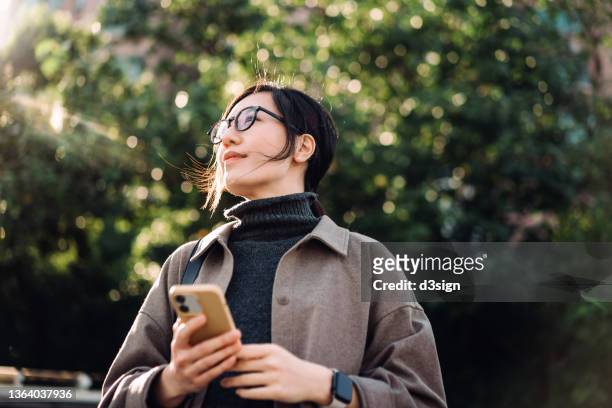low angle portrait of young asian woman using smartphone in park against sunlight, looking up with smile, enjoying the fresh air and gentle breeze in the nature. lifestyle and technology - business air travel photos et images de collection