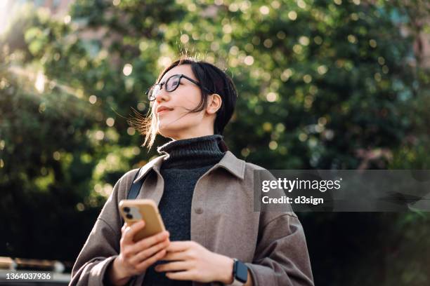 low angle portrait of young asian woman using smartphone in park against sunlight, looking up with smile, enjoying the fresh air and gentle breeze in the nature. lifestyle and technology - business smile stockfoto's en -beelden