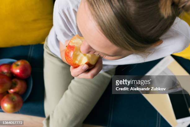 a red apple in the hands of a woman who is sitting on a bright-colored sofa at home. the girl eats a fruit, bites off a piece of juicy apple. cultivation of organic farm products. the concept of vegetarian, vegan, raw food and diet. - pomme croquée photos et images de collection