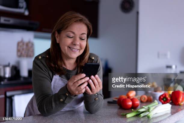 alegre mujer latinoamericana enviando mensajes de texto en su teléfono inteligente mientras se apoya en el mostrador de la cocina antes de cocinar una comida en casa - mature latin women fotografías e imágenes de stock