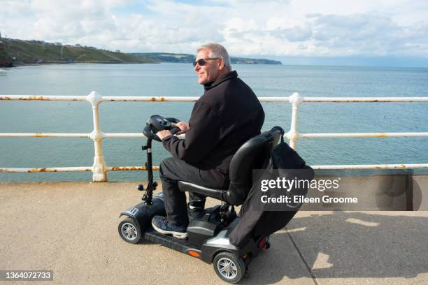 middle aged male sits on his mobility scooter looking at the sea from the promenade at new quay in wales hoping to see dolphins or other wildlife, enjoying the freedom his scooter gives him to be able to get out and about unaided. - mobility scooters stock pictures, royalty-free photos & images