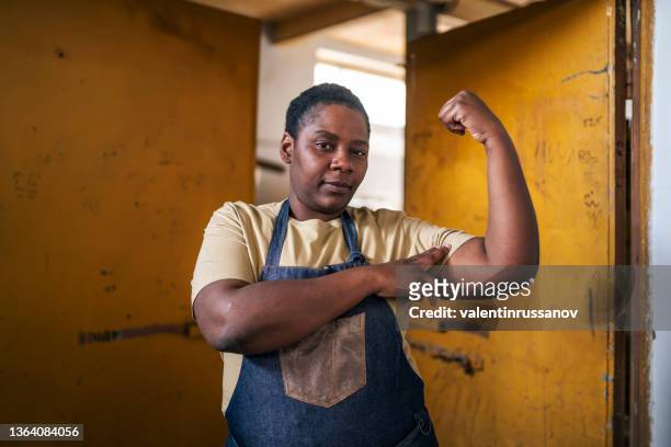 porträt einer selbstbewussten afro-frau, die arbeitskleidung trägt und stark aussieht, während sie ihren bizeps beugt - bürgerrecht stock-fotos und bilder