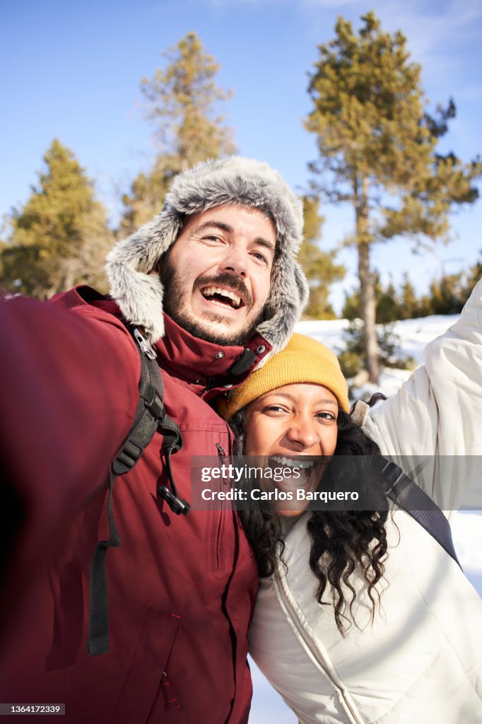 Vertical pohoto of an interracial young couple taking a selfie in a snowy landscape.