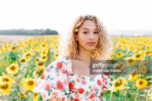 a young woman in a floral dress looking at the camera in a field of sunflowers. - floral pattern dress stockfoto's en -beelden