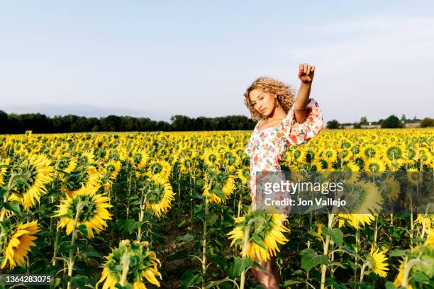 a young woman standing in a field of sunflowers raising one arm while looking at the camera. - happy sunflower stock pictures, royalty-free photos & images