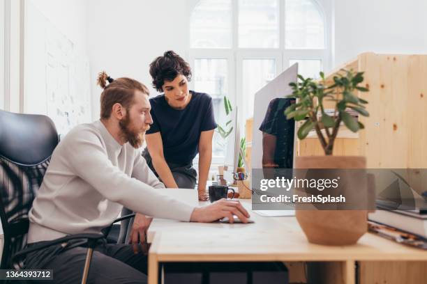 a hardworking man and his female colleague looking at the screen of his computer and analyzing some statistics - desk with green space view stock pictures, royalty-free photos & images