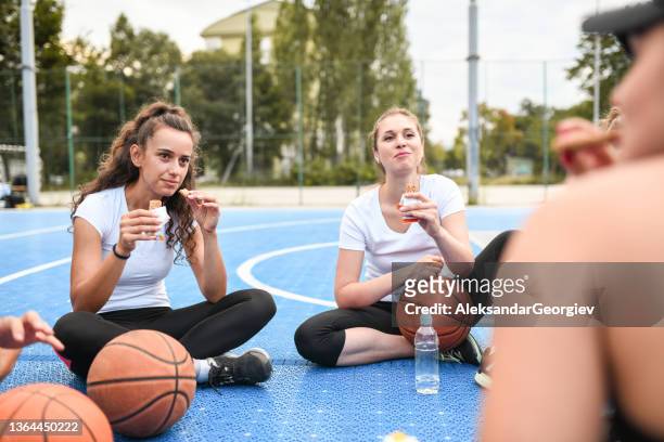 cute females eating protein bar before basketball training on court - maillot de sport stockfoto's en -beelden