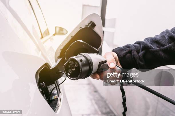 detail of a hand plugging the cord into an electric car, to charge the battery in the garage outside a home. concept of electric car charging, renewable energy, sustainability and transport. - opladen stockfoto's en -beelden