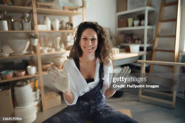 mid adult woman, showing her dirty hands from making pottery on a pottery wheel and looking at camera in art studio. - pottery wheel stock pictures, royalty-free photos & images