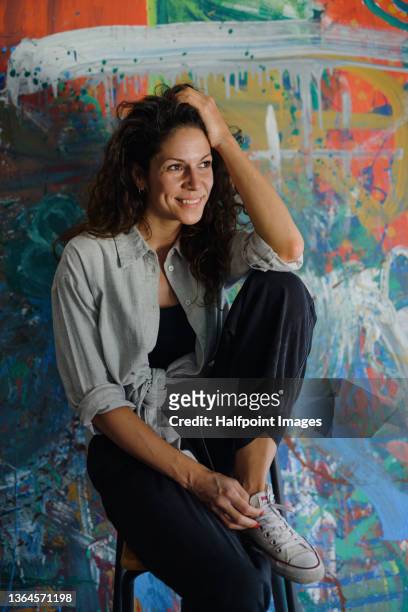 mid adult woman artist sitting in front of her painting and looking away. - art and craft stock pictures, royalty-free photos & images