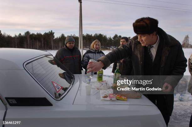 Unknown Ukrainian man in sheepskin coat and fur hat, pouring vodka shots on the boot of his car, to drink with a picnic of meat and bread, with...