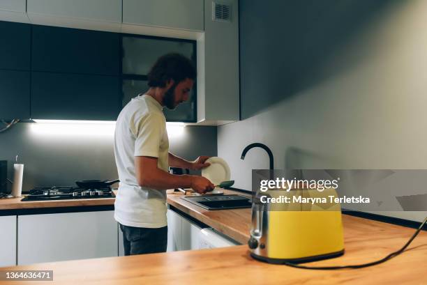 a young guy washes the dishes in the kitchen. morning. yellow toaster. bearded guy. modern cuisine. house cleaning. - toaster fotografías e imágenes de stock