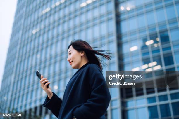 low angle portrait of successful young asian businesswoman using smartphone in city, standing against contemporary corporate skyscrapers in financial district. female leadership, girl power, business on the go concept - low confidence stock pictures, royalty-free photos & images