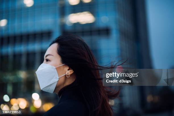 side profile of confident young asian businesswoman with protective face mask looking ahead, standing against illuminated contemporary corporate skyscrapers in financial district in the city. female leadership, girl power, business on the go concept - china coronavirus stock pictures, royalty-free photos & images