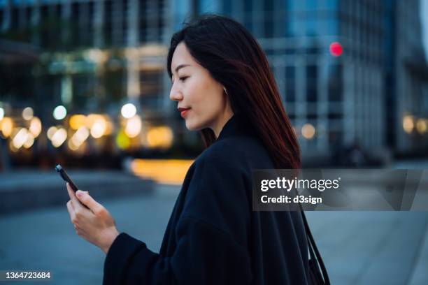 independence and successful young asian businesswoman managing finance and investment with online banking on smartphone on the go, with illuminated corporate skyscrapers in background. smart banking anytime anywhere with technology concept - frank rich stock pictures, royalty-free photos & images