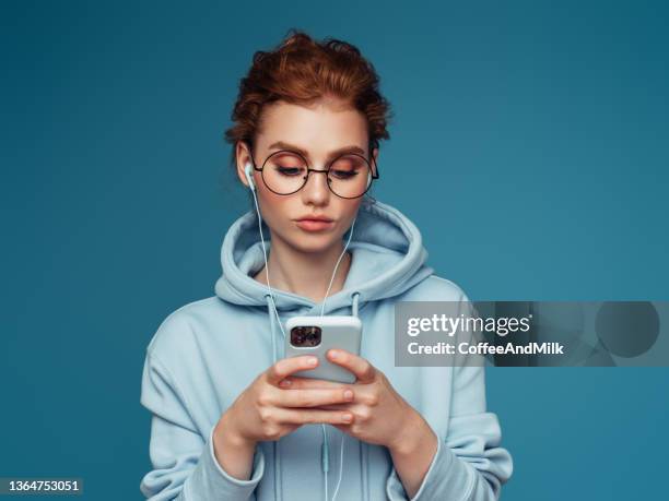 close-up portrait of a young pretty girl in a blue hoodie using smart phone - hoodie headphones stock pictures, royalty-free photos & images