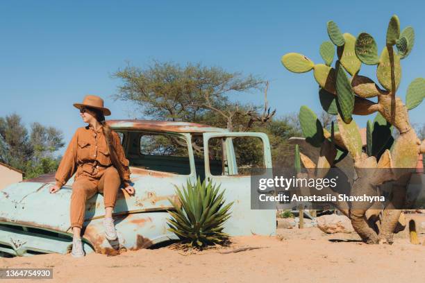 weibliche reisende mit hut sitzt auf dem alten verlassenen auto mitten in der wüste in namibia - wildunfall stock-fotos und bilder