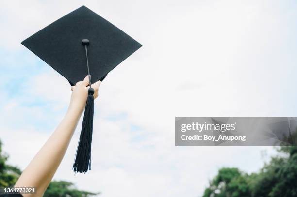 student hand celebrating graduation by holding and throwing cap against the sky. - traje academico imagens e fotografias de stock