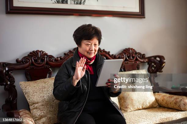 an old woman of east asian descent is chatting online on a tablet computer on a chinese chair in the living room - china middle class stock pictures, royalty-free photos & images