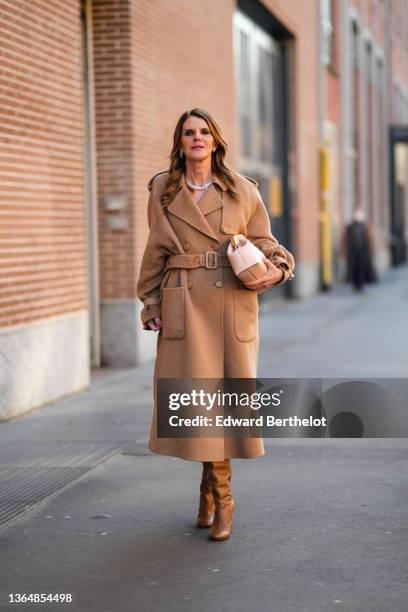 Anna Dello Russo wears a bejeweled necklace, a beige brown wool long winter double breasted trench coat with a large belt, a pastel pale pink Fendi...