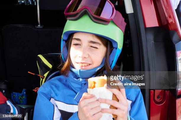 close-up of front view of a girl dressed in winter clothes sitting in the trunk of the car looking away has a snack while taking a break - sports car photos et images de collection