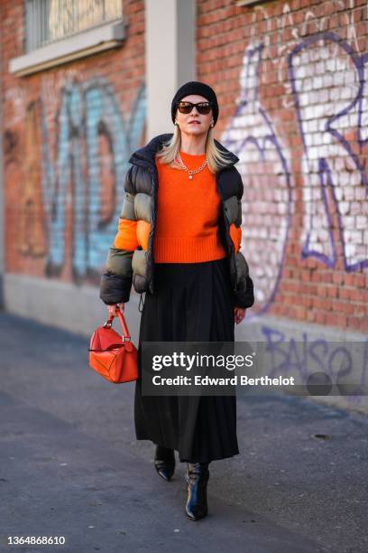Guest wears a beanie hat, a necklace, a necklace, an orange ribbed wool pullover, a khaki black and orange striped winter puffer jacket, a black...