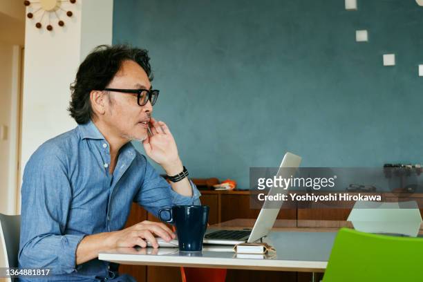senior male teleworking in his living room at home, view from the side of his laptop. - japanese old man stock-fotos und bilder