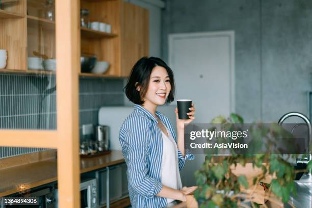 jeune femme asiatique détendue dégustant une tasse de café le matin près du comptoir de la cuisine à la maison, rêvant tout en détournant le regard - asian drink photos et images de collection