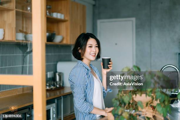 relaxed young asian woman enjoying a cup of morning coffee by the kitchen counter at home, day dreaming while looking away - zen-like stock pictures, royalty-free photos & images