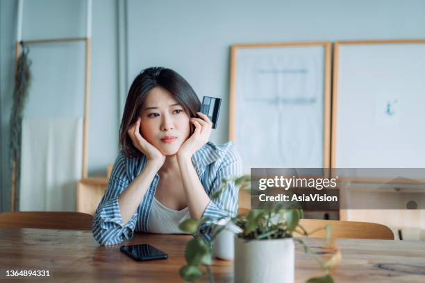 young asian woman having problems with her credit card and looking worried. financial issues, banking, finance, credit card fraud - thinking for investment stock pictures, royalty-free photos & images