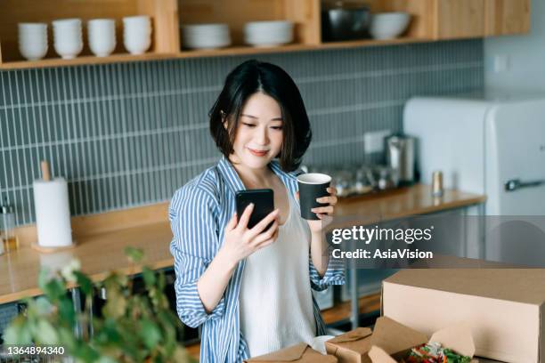 beautiful smiling young asian woman standing by the kitchen counter, using smartphone while enjoying assorted home delivery takeaway food at home. eating at home concept - customer experience stock pictures, royalty-free photos & images