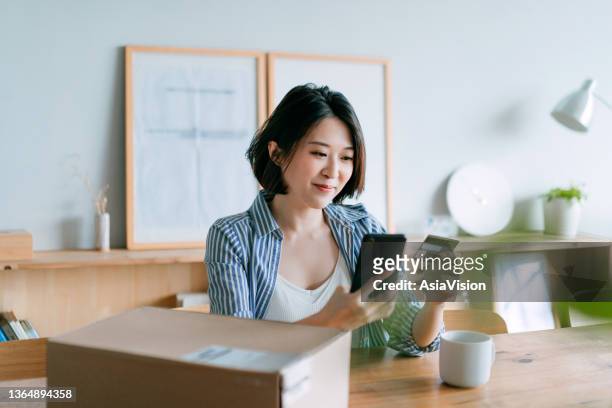cheerful young asian woman shopping online with smartphone and making mobile payment with credit card on hand, receiving a parcel by home delivery service. technology makes life so much easier - asian credit card imagens e fotografias de stock